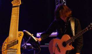 Eine Mann spielt eine akustische Gitarre auf einer Bühne, daneben steht ein großes, mehrsaitiges Saiteninstrument Namens Koyabu-Symmetric-Board.