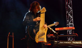 Eine Person spielt ein großes, ungewöhnliches Saiteninstrument (Koyabu-Symmetric-Board) auf einer Bühne, umgeben von Musikgeräten und Scheinwerfern.
