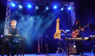 Zwei Personen auf einer Bühne mit blauer Beleuchtung, einer spielt Keyboard und Gitarre; im Hintergrund sind weitere Instrumente sichtbar.