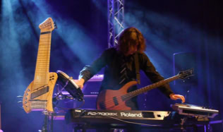 Eine Person spielt auf einer Bühne ein E-Bass und ein Keyboard, mit einem weiteren Saiteninstrument (Koyabu-Symmetric-Board) auf einem Ständer daneben. Im Hintergrund sind Lichteffekte sichtbar.