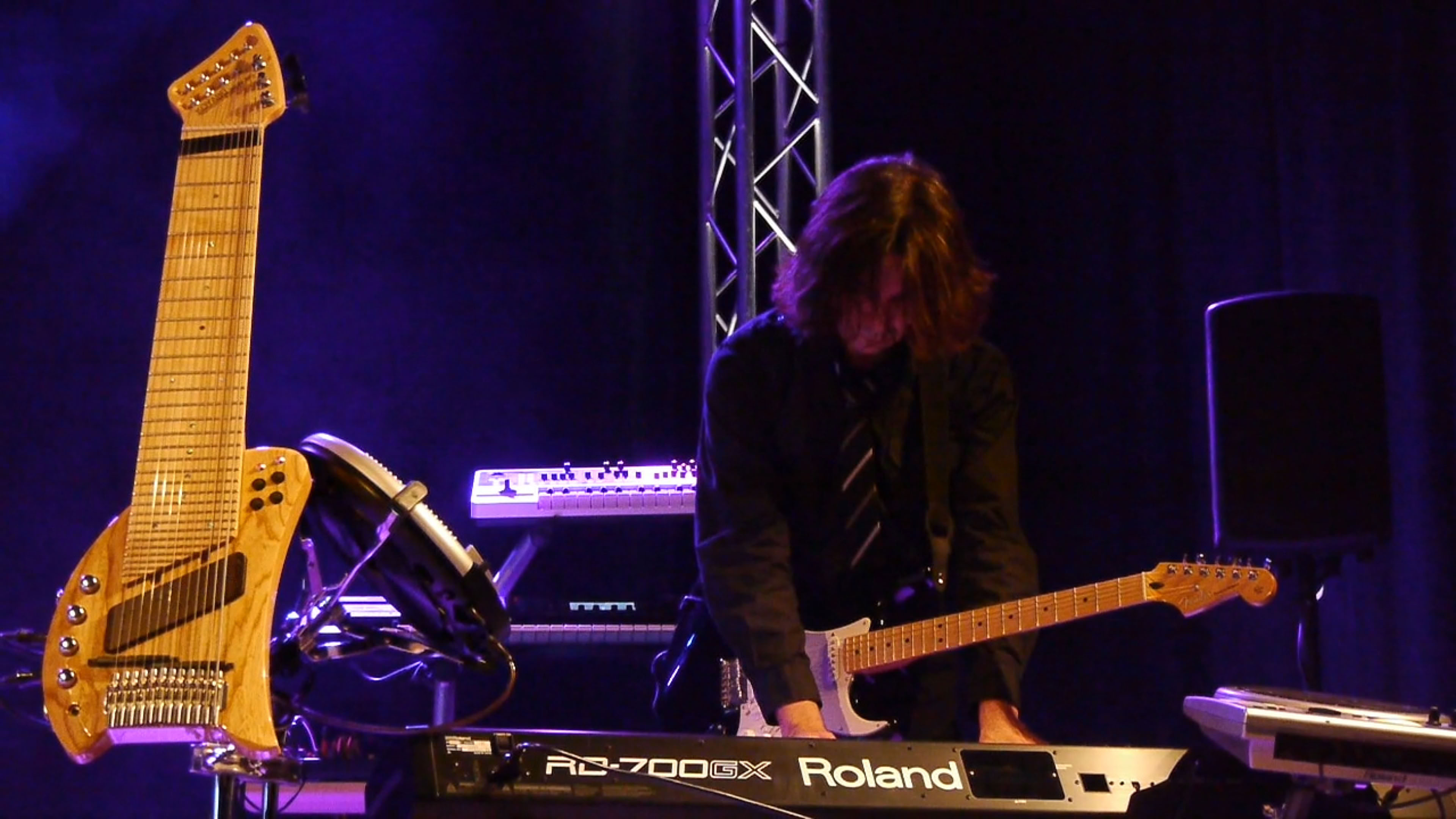 Person spielt Keyboard und Gitarre auf einer Bühne mit Lichteffekten im Hintergrund. Ein weiteres Saiteninstrument (Koyabu-Symmetric-Board) steht links.