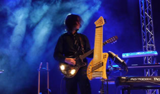 Musiker auf der Bühne mit einer Gitarre und einem ungewöhnlichen Saiteninstrument (Koyabu-Symmetric-Board), umgeben von blauer Bühnenbeleuchtung und Nebel.