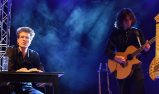 Zwei Personen auf einer Bühne, eine sitzt an einem Tisch mit einem Buch, die andere spielt Gitarre; blauer Hintergrund mit Bühnenbeleuchtung.