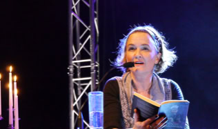 Eine Person mit Mikrofon hält ein Buch in der Hand, im Hintergrund sind Kerzen und eine Bühnenstruktur zu sehen.