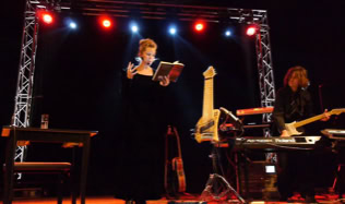 Eine Frau steht auf einer Bühne und liest aus einem Buch, während ein Mann ein Musikinstrument spielt. Im Hintergrund sind bunte Lichter und Bühnenequipment zu sehen.