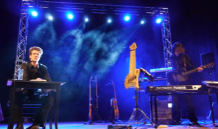 Ein Sprecher und ein Musiker auf einer Bühne mit blauer Beleuchtung, der Musiker spielt Keyboard und Gitarre; im Hintergrund sind weitere Instrumente sichtbar.