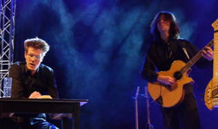 Zwei Personen auf einer Bühne, eine sitzt an einem Tisch, die andere spielt Gitarre; blaues Bühnenlicht im Hintergrund.