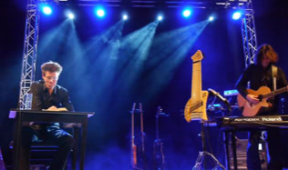 Ein Sprecher und ein Musiker auf einer Bühne mit blauer Beleuchtung, der Musiker spielt Keyboard und Gitarre.