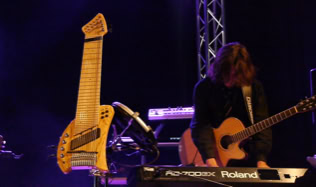 Ein Mann spielt Gitarre und steht vor einem Keyboard auf einer Bühne, daneben steht ein ungewöhnliches Saiteninstrument (Koyabu-Symmetric-Board).