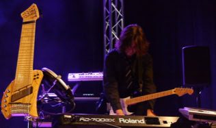 Person spielt E-Gitarre und Keyboard auf einer Bühne, im Hintergrund sind Lichteffekte und ein weiteres Musikinstrument (Koyabu-Symmetric-Board) zu sehen.