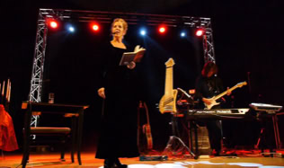 Eine Frau steht auf einer Bühne und liest aus einem Buch, während ein Musiker Gitarre spielt. Im Hintergrund sind bunte Lichter und Bühnenequipment zu sehen.