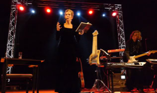 Eine Frau steht auf einer Bühne und hält ein Buch, während sie spricht. Im Hintergrund sind Lichter und ein Musiker mit einem Keyboard zu sehen.